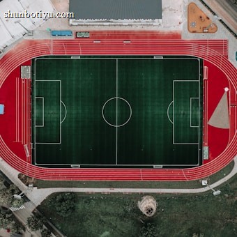 An aerial view of a large sports complex featuring a central green football field surrounded by a red running track. Adjacent to the track, there is a parking area, buildings, and pathways. Nearby, there are additional sports courts in different colors, including a blue swimming pool and orange basketball courts.