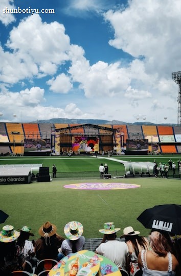 A sports stadium with a large stage set up on the field, hosting an outdoor event. Spectators wearing colorful hats sit in the foreground. The seating areas display vibrant colors, and a clear blue sky with scattered clouds is visible. Large screens display graphics related to the event.